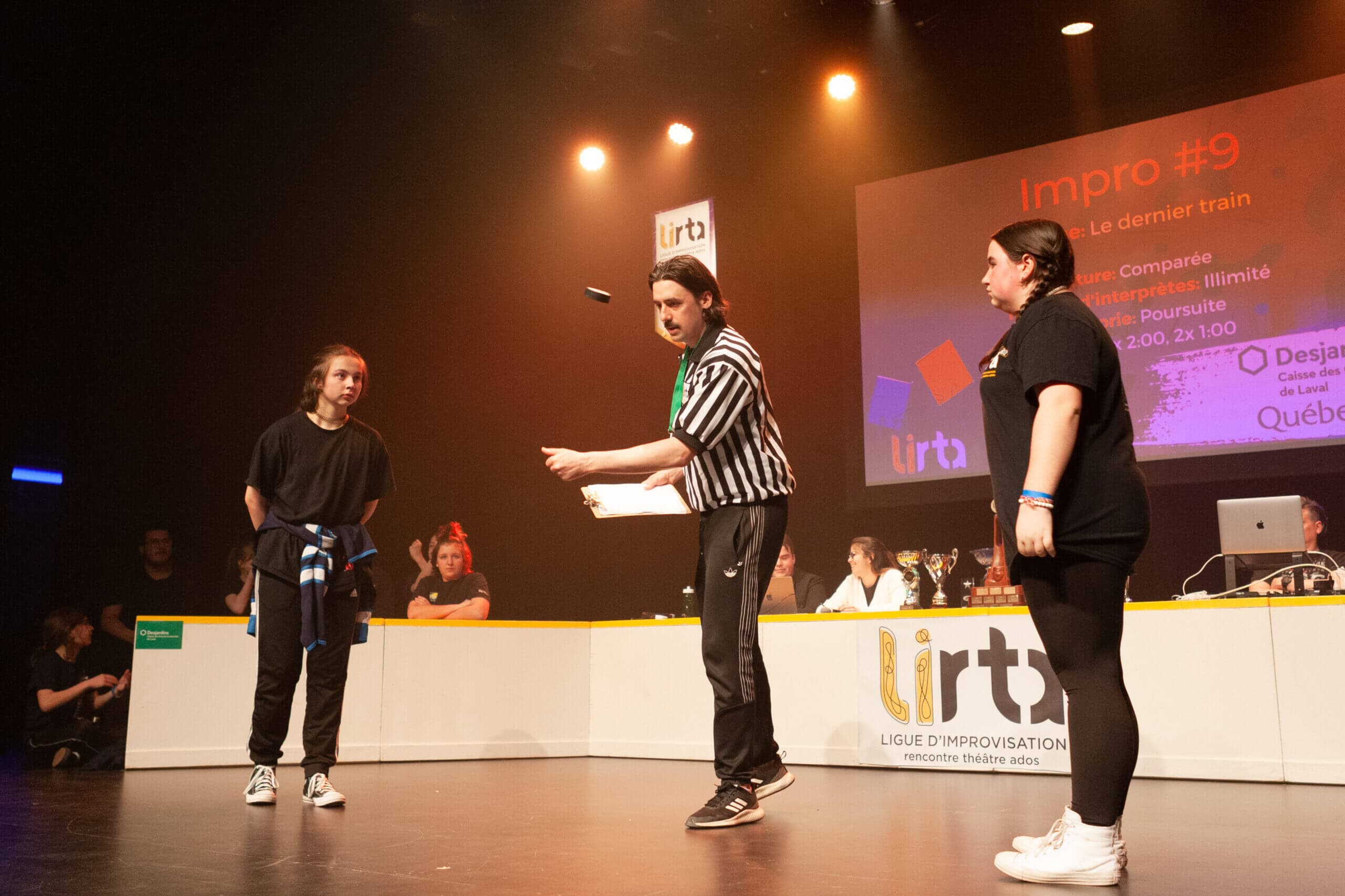On stage, a referee in a striped shirt tosses a token in the air between two participants dressed in black, in front of a screen displaying “Impro #9.”