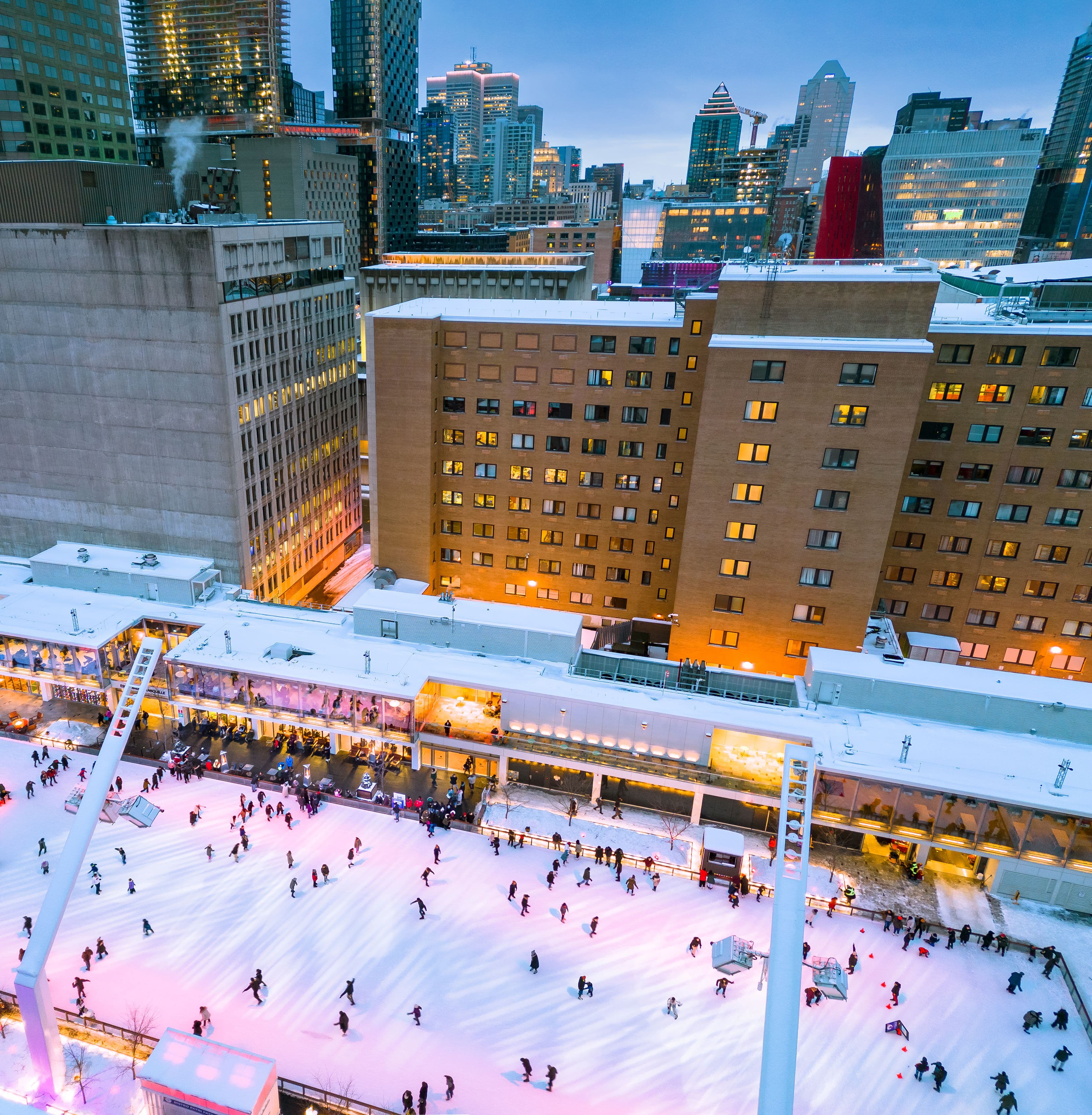 Aerial view of an outdoor ice rink glowing with pink lights, lively with skaters, at the foot of tall buildings in a snowy downtown.