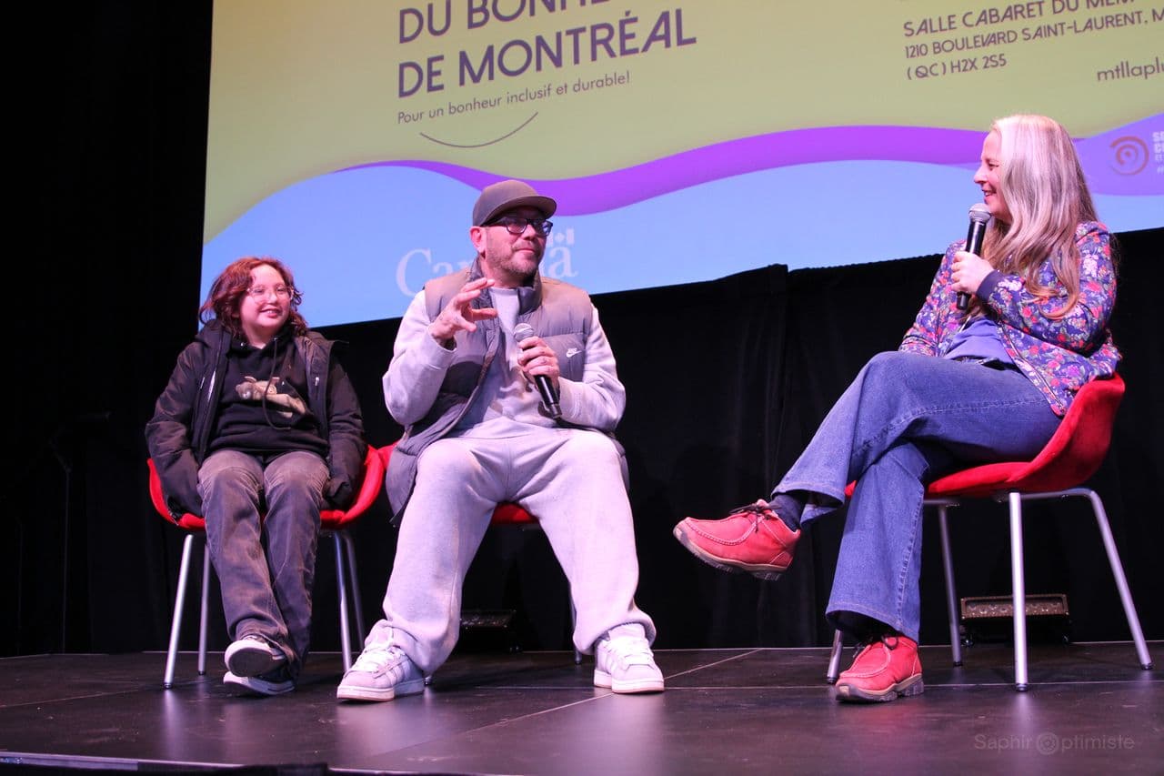 Three people sitting on red chairs, talking on a stage.