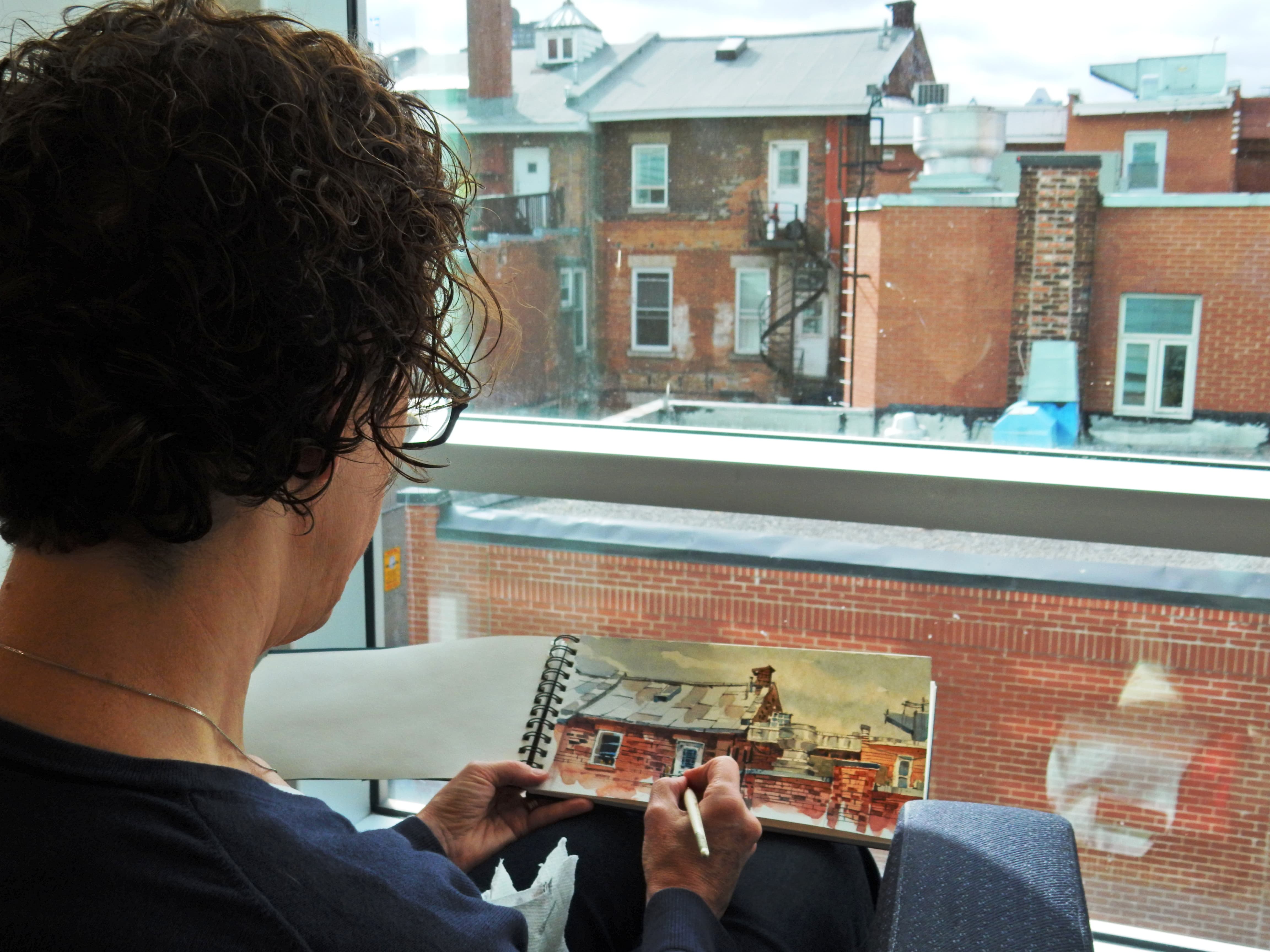 A person sketches red brick buildings in a notebook while sitting by a large window overlooking rooftops and urban facades.