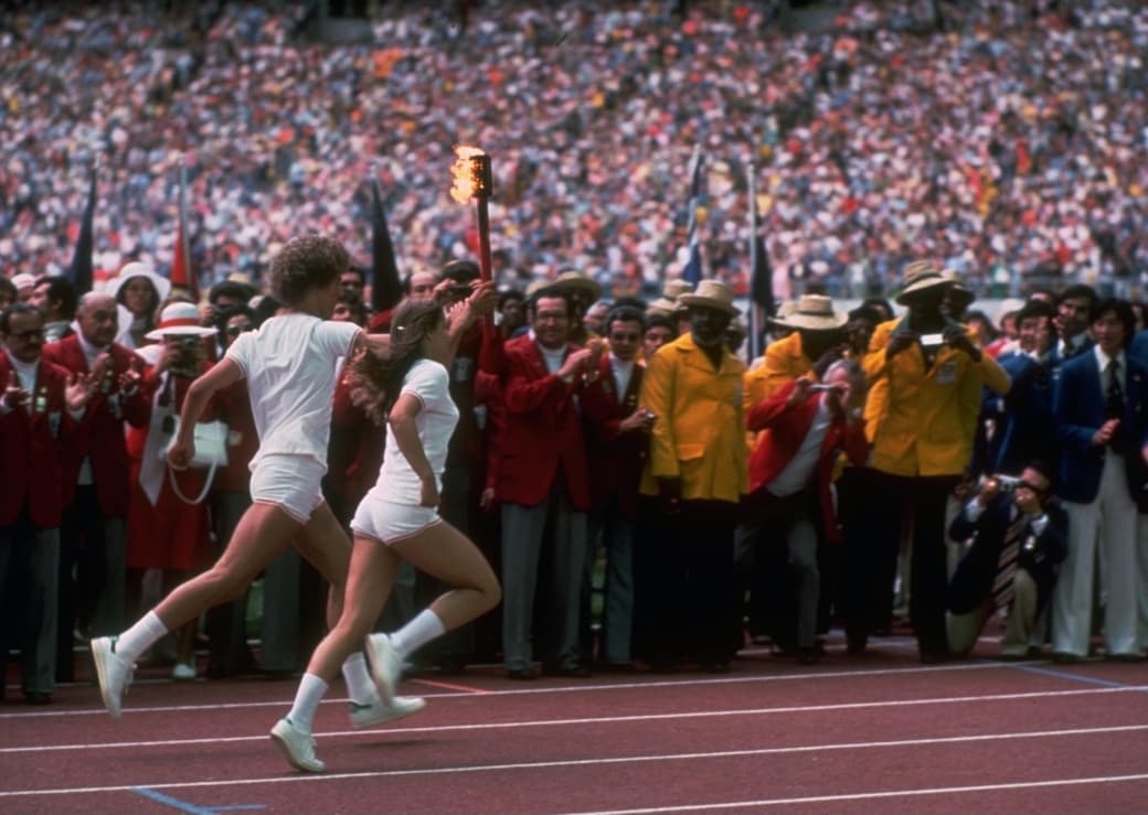 A man and a woman running on the Olympic Stadium track and carrying the Olympic torch in 1976.