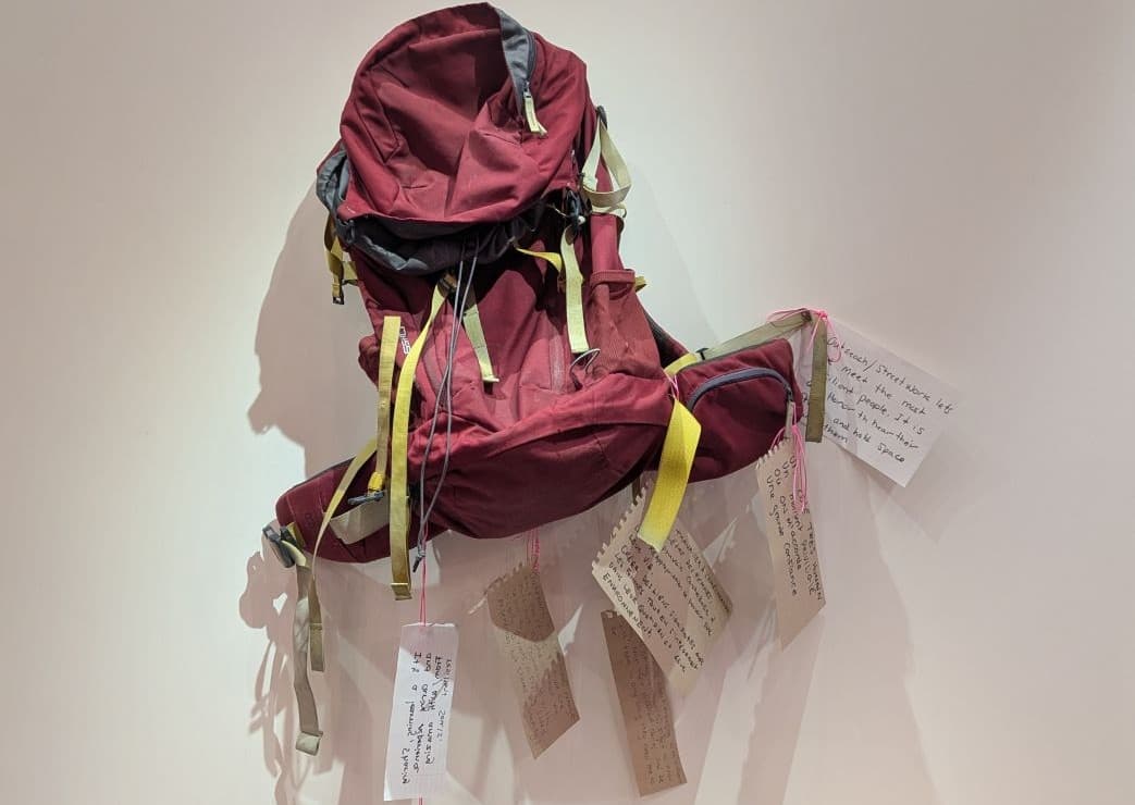 Un sac à dos bordeaux accroché à un mur blanc, décoré de plusieurs étiquettes manuscrites suspendues à ses sangles.